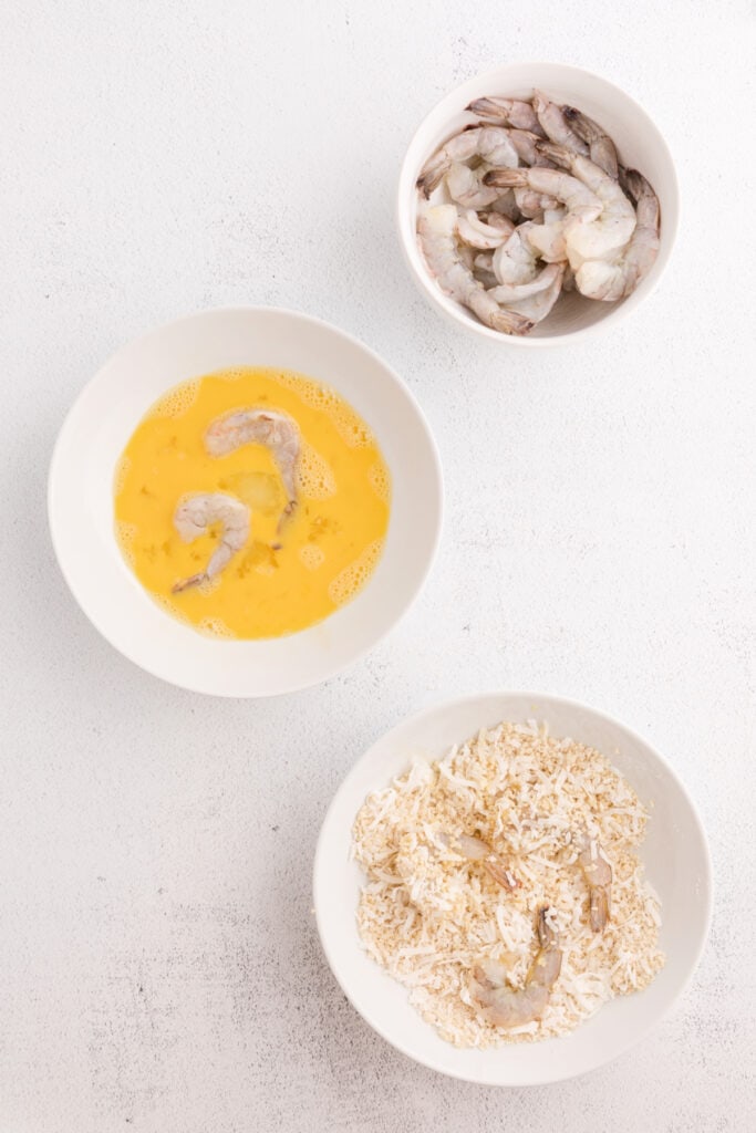 Shrimp dipped in beaten eggs and coated in a coconut and breadcrumb mixture in separate bowls.
