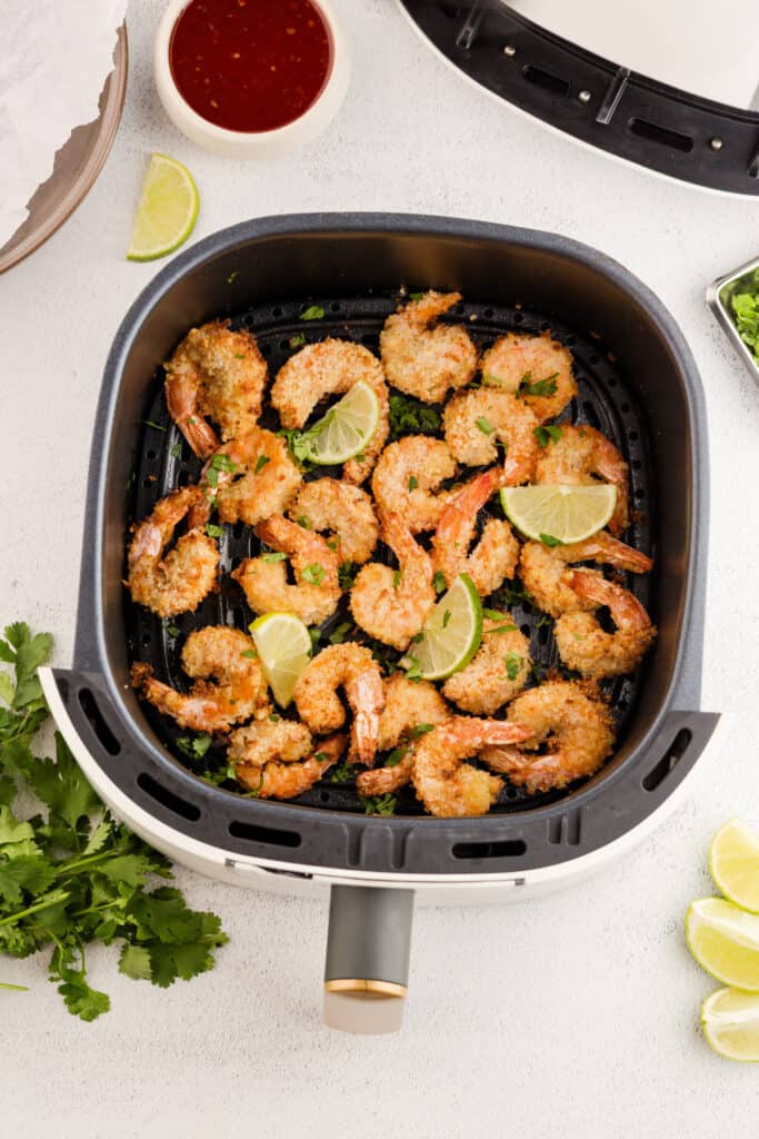 Cooked coconut shrimp in an air fryer basket with lime wedges and chopped cilantro.