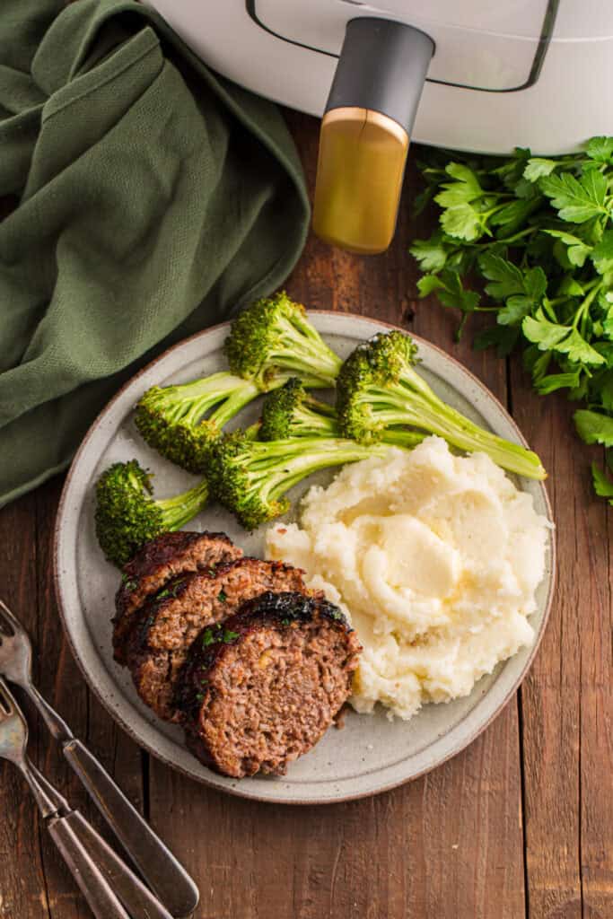 Plate with sliced meatloaf, mashed potatoes, and broccoli next to an air fryer on a wooden table.