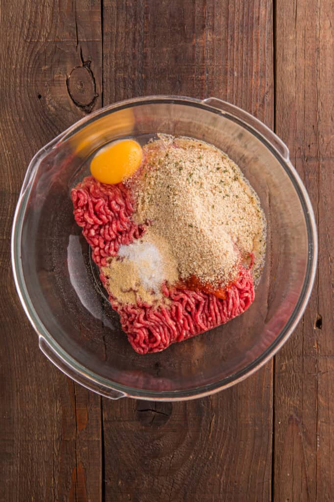 Ground beef, egg, breadcrumbs, chopped onion, ketchup, and seasonings in a clear glass mixing bowl on a wooden surface.
