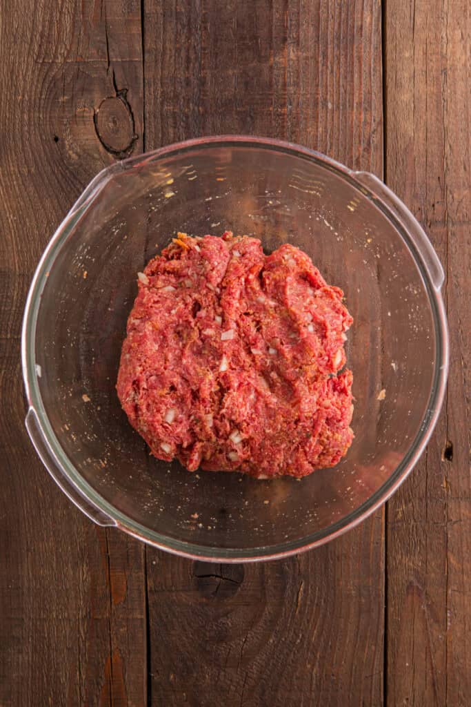 Mixed raw meatloaf mixture with visible chopped onion in a clear glass bowl on a wooden table.