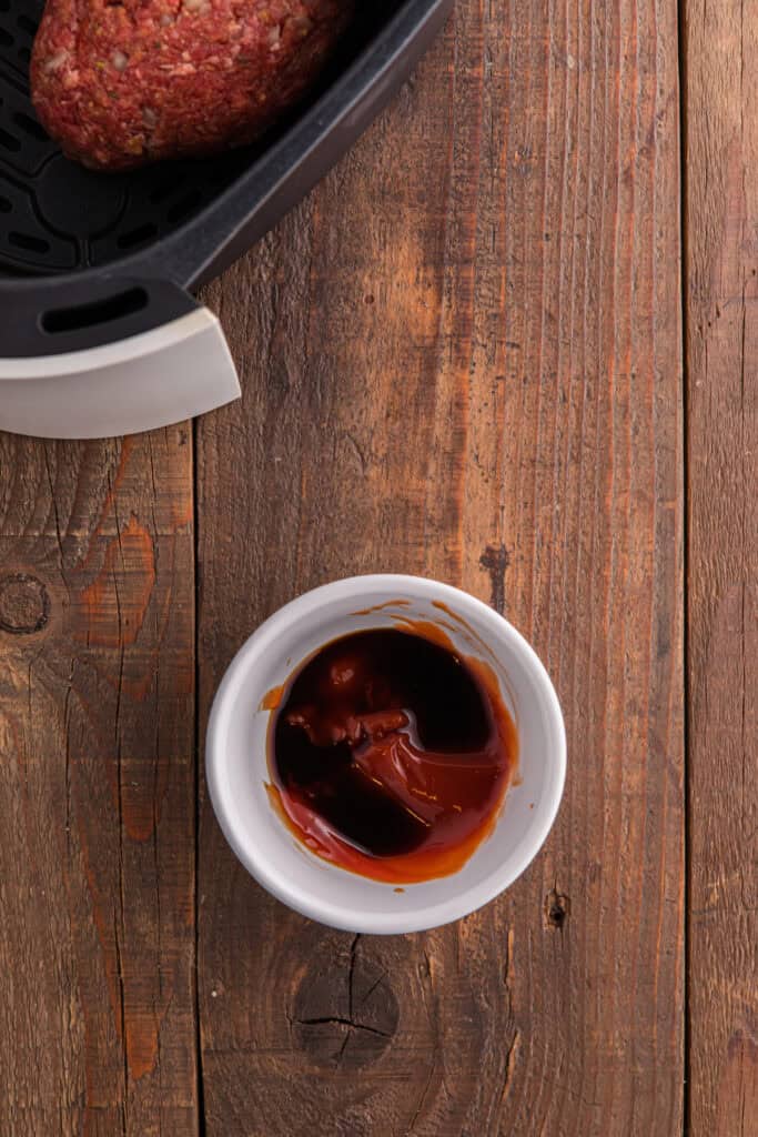 Small white bowl with ketchup and Worcestershire sauce mixture on a wooden surface next to an air fryer basket.