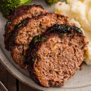 Close up of sliced glazed meatloaf on a plate with mashed potatoes and broccoli.