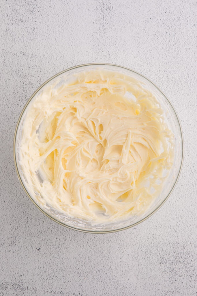 Glass bowl filled with smooth cream cheese frosting on a light textured surface