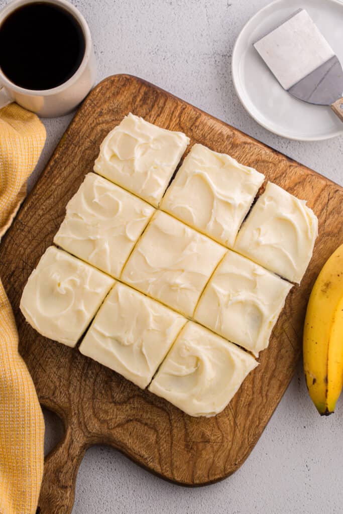 Frosted banana brownies cut into squares on a wooden cutting board with a cup of coffee and bananas nearby