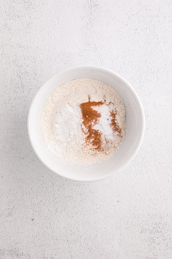 White bowl filled with all-purpose flour, baking soda, baking powder, salt, and ground cinnamon on a light textured surface