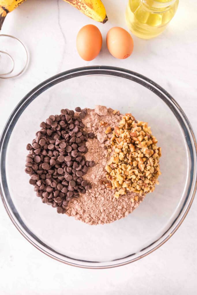Glass mixing bowl with brownie mix, chocolate chips, and chopped walnuts on a marble countertop.