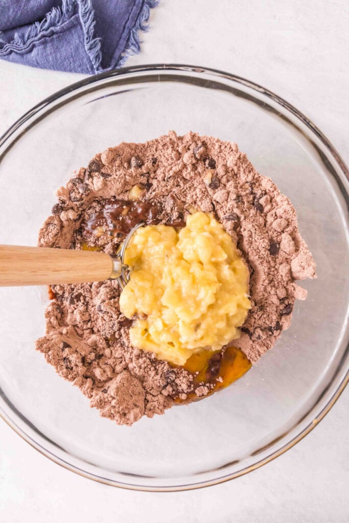 Mixing bowl with brownie mix, chocolate chips, walnuts, eggs, oil, and mashed bananas being added.