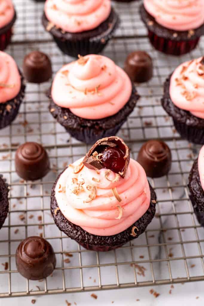 Overhead view of frosted chocolate cupcakes with pink frosting and cherry cordial candies arranged on a wire rack