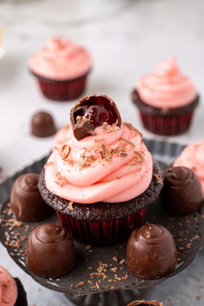 Close up view of a chocolate cupcake topped with pink frosting, chocolate shavings, and a halved cherry cordial candy, surrounded by whole cherry cordial candies