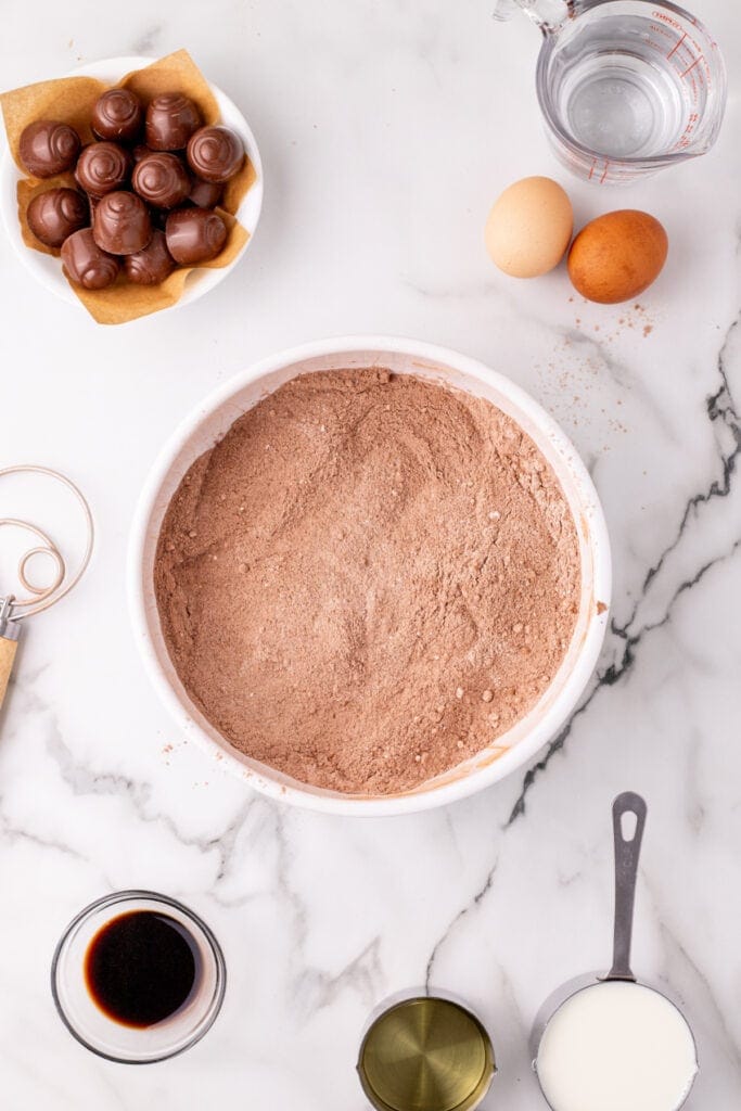 Overhead view of a bowl with chocolate cake dry ingredients fully mixed together, surrounded by eggs, milk, oil, vanilla, water, and cherry cordial candies