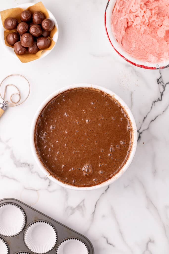 Overhead view of a bowl filled with smooth chocolate cake batter, with cupcake liners and mixing tools nearby