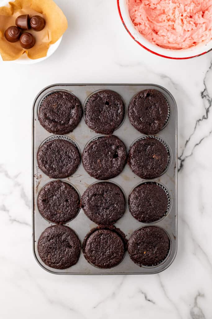 Overhead view of baked chocolate cupcakes cooling in a muffin pan next to a bowl of pink frosting and cherry cordial candies