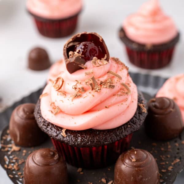 Close up view of a chocolate cupcake with pink frosting and a halved cherry cordial candy on top, with other frosted cupcakes blurred in the background
