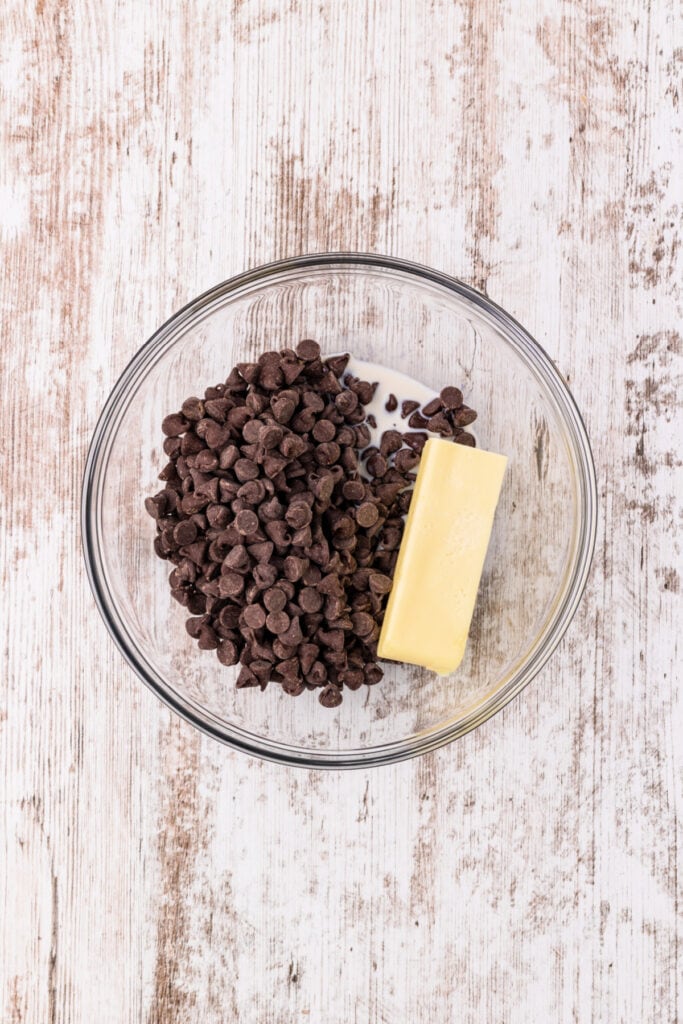 Glass bowl with semisweet chocolate chips and a stick of butter on a white wood surface.