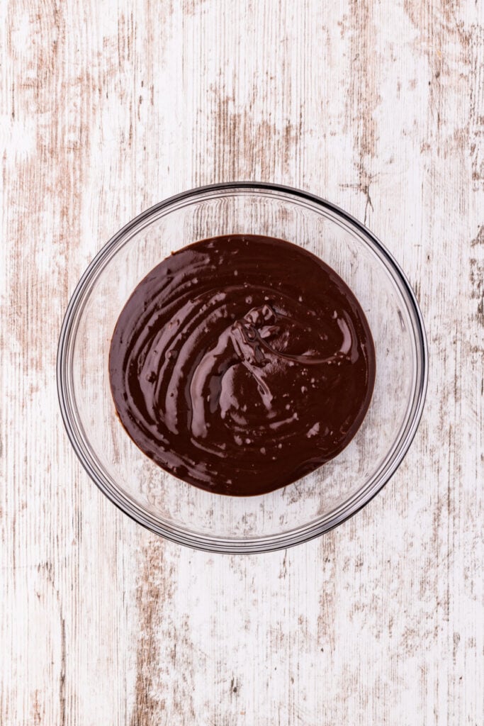 Melted chocolate mixture in a clear glass bowl viewed from above on a white wood surface.
