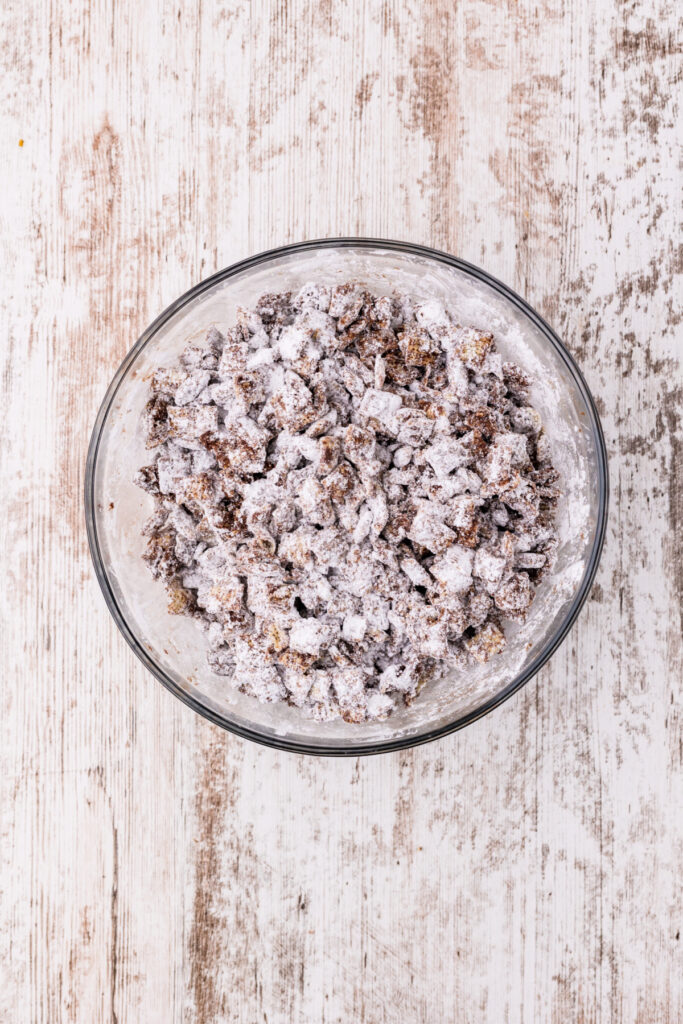 Chocolate coated Chex cereal covered in powdered sugar in a clear glass bowl viewed from above.