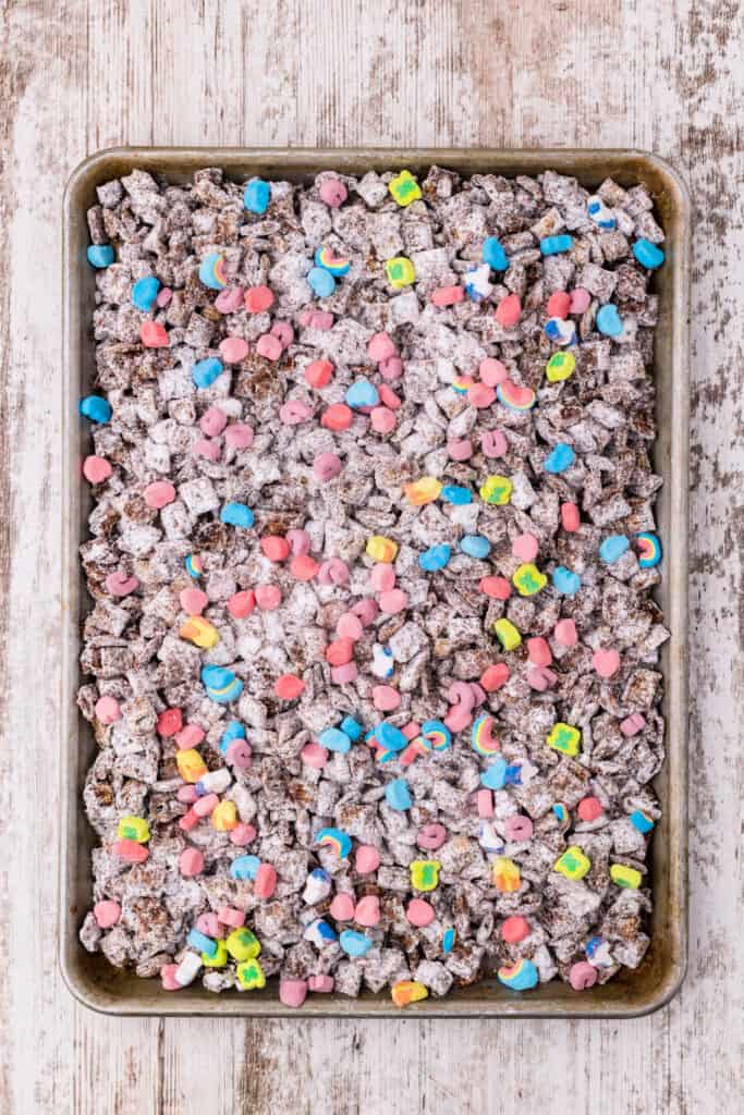 Powdered sugar coated Chex cereal in a metal baking sheet topped with colorful Lucky Charms marshmallows viewed from above.