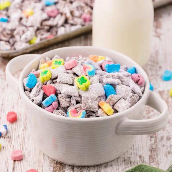 White handled bowl filled with powdered sugar coated Chex cereal and colorful marshmallows with a glass bottle of milk in the background on a white wood surface.