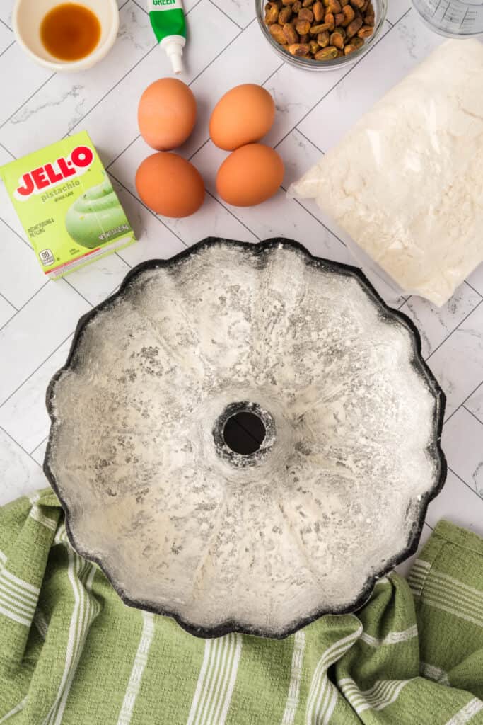 floured bundt pan on a countertop surrounded by eggs, pistachio pudding mix, pistachios, flour, and food coloring