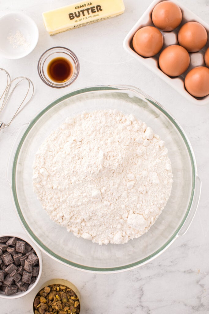 Overhead view of a glass mixing bowl filled with flour surrounded by butter, eggs, vanilla, chocolate chunks, chopped pistachios, and a whisk on a white surface.