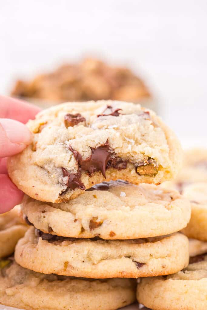 Hand holding a broken pistachio chocolate chunk cookie with melted chocolate center over a stack of cookies.