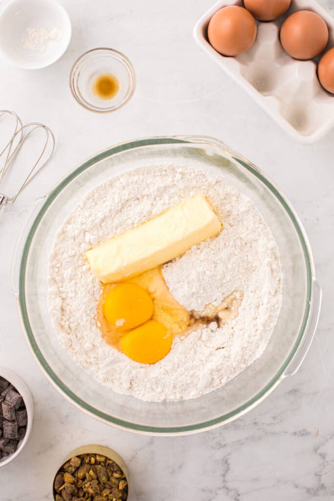 Overhead view of flour in a glass bowl with softened butter, two eggs, and vanilla added, with baking tools and ingredients around the bowl.