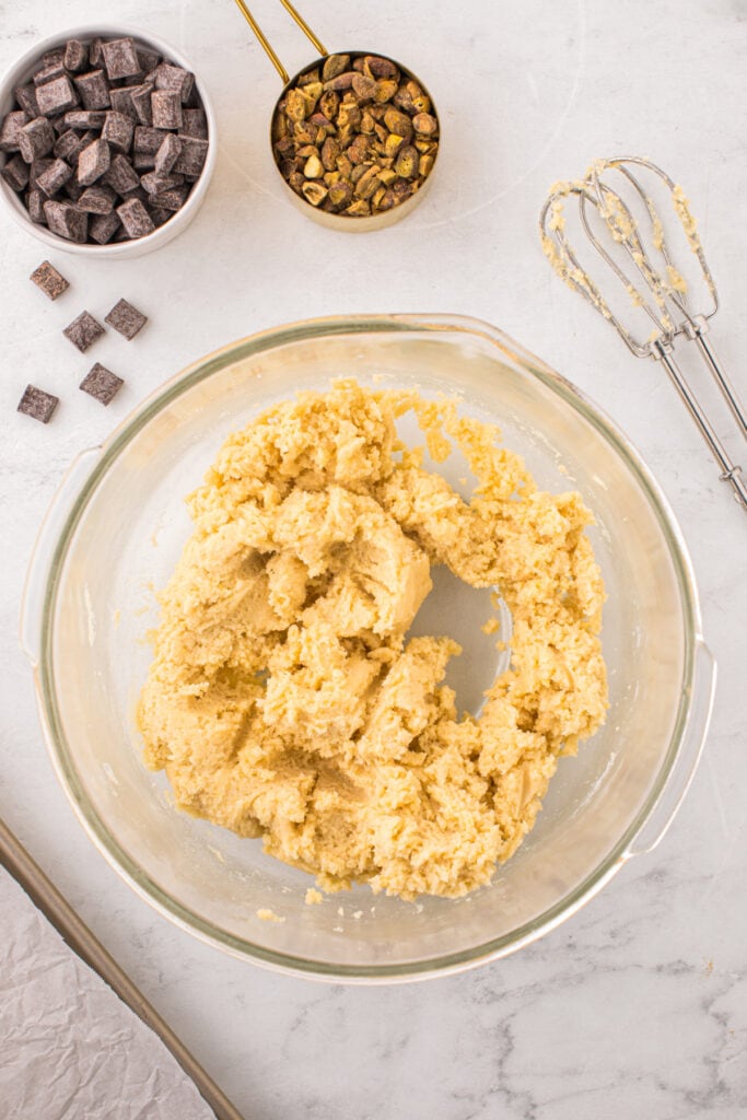 Overhead view of thick cookie dough mixed in a glass bowl with chocolate chunks and chopped pistachios nearby.