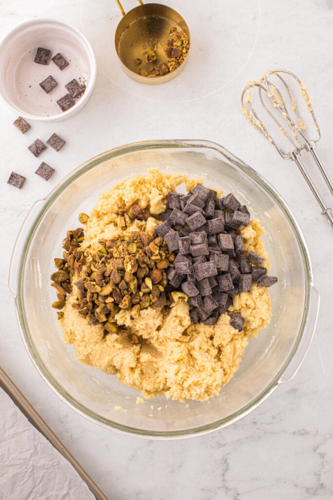 Overhead view of cookie dough in a glass bowl topped with chopped pistachios and chocolate chunks before mixing.