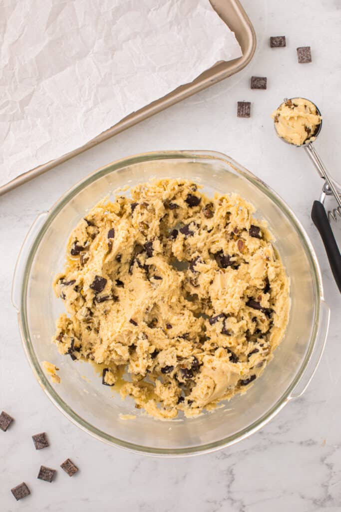 Overhead view of fully mixed pistachio chocolate chunk cookie dough in a glass bowl with a cookie scoop nearby.