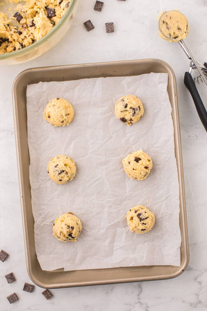 Overhead view of six scooped balls of pistachio chocolate chip cookie dough spaced on a parchment lined baking sheet.