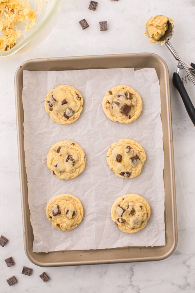Overhead view of six baked pistachio chocolate chunk cookies on a parchment lined baking sheet.