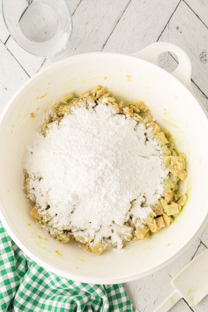 Powdered sugar added on top of green coated cereal in large white mixing bowl