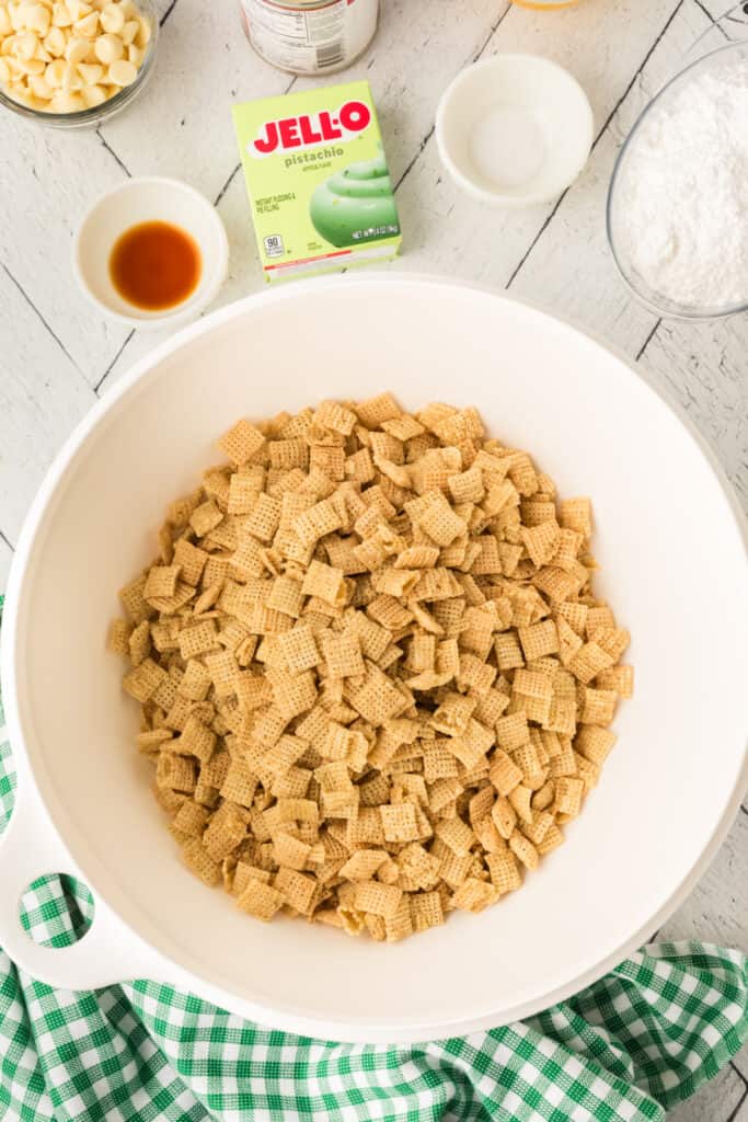 Large white mixing bowl filled with Rice Chex cereal on a white wood surface with pistachio pudding mix box, powdered sugar, vanilla, and salt nearby