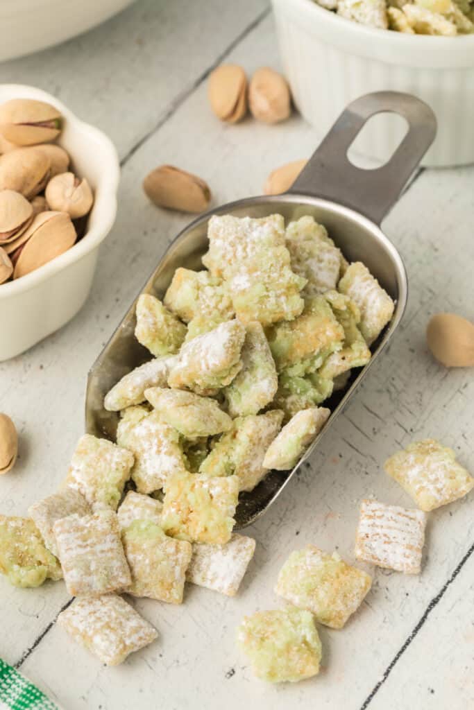 Metal scoop filled with pistachio muddy buddies on a light surface with pistachios and white ramekins in the background