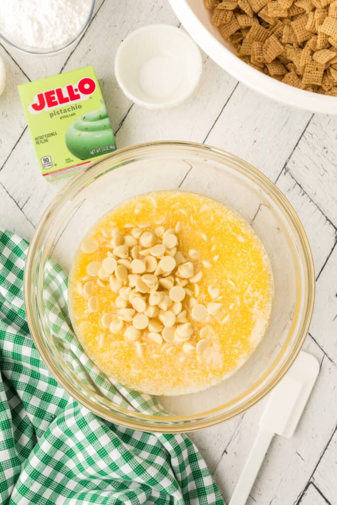 Glass bowl with melted butter, sweetened condensed milk, and white chocolate chips before stirring, with pistachio pudding mix box in background