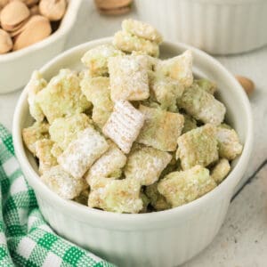 Square cropped image of pistachio muddy buddies in a white ramekin dusted with powdered sugar