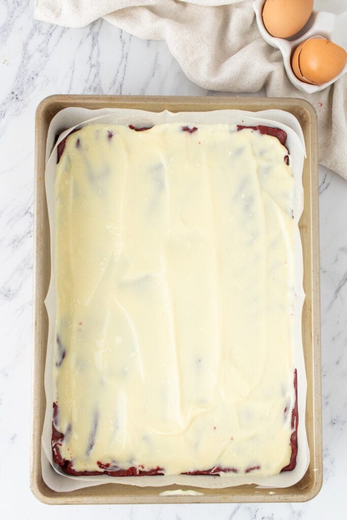 Cream cheese layer spread over red velvet batter in a parchment lined 9x13-inch baking pan