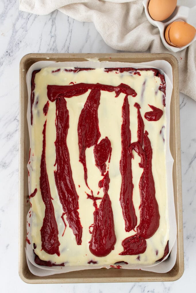 Red velvet batter spooned in lines over cream cheese layer in a parchment lined 9x13-inch baking pan