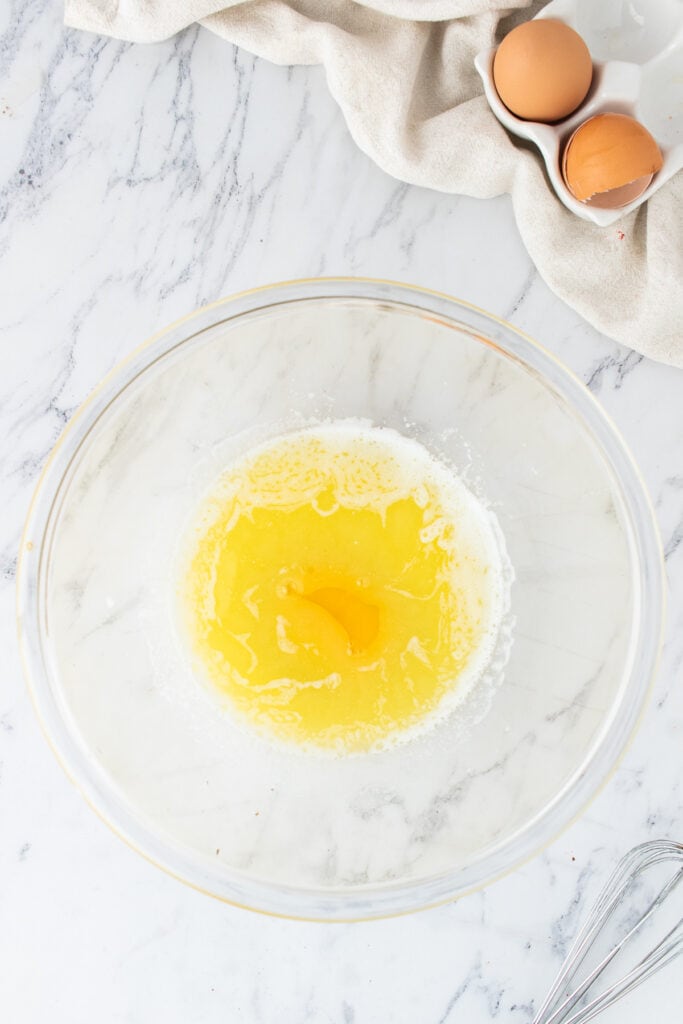 Glass mixing bowl with melted butter and a cracked egg on a marble surface with eggs and cloth nearby