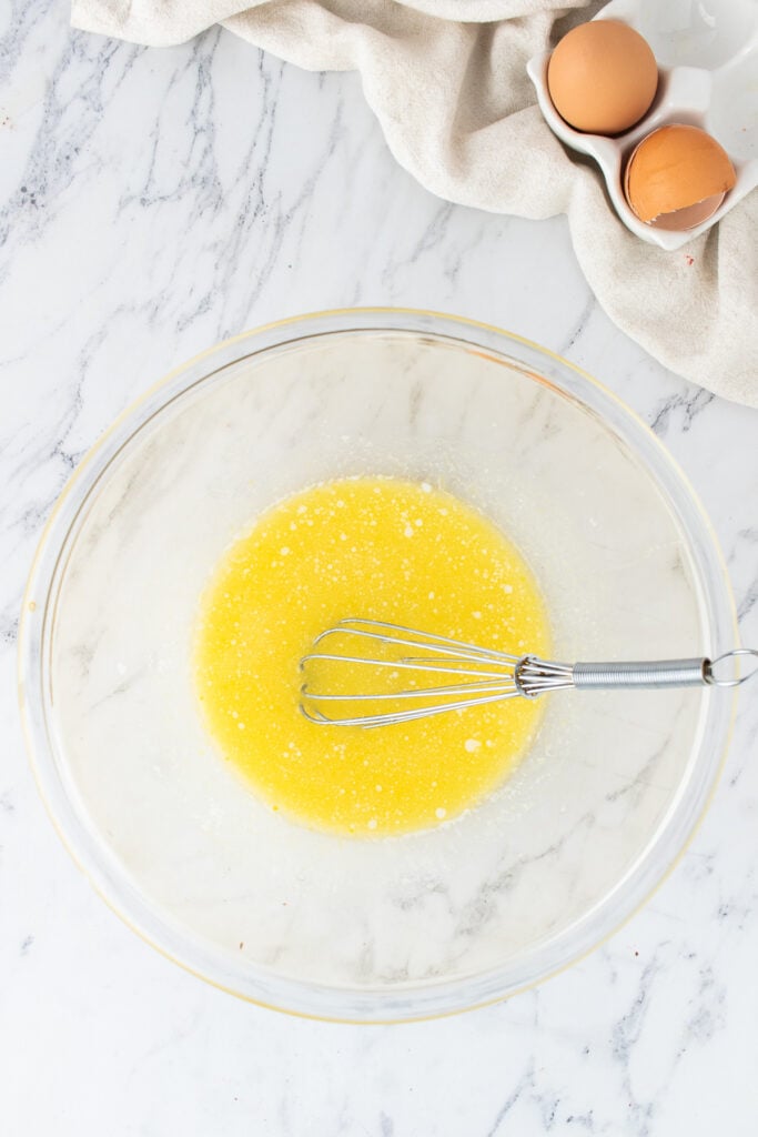 Glass mixing bowl with melted butter mixture and a whisk resting inside on a marble surface