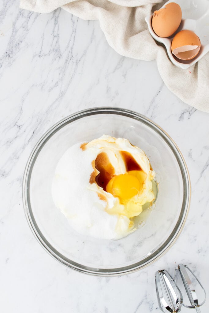 Glass bowl with cream cheese, sugar, egg, and vanilla before mixing on a marble surface