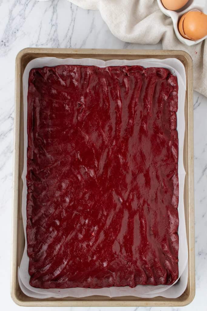 Red velvet brownie batter spread evenly in a parchment lined 9x13-inch baking pan