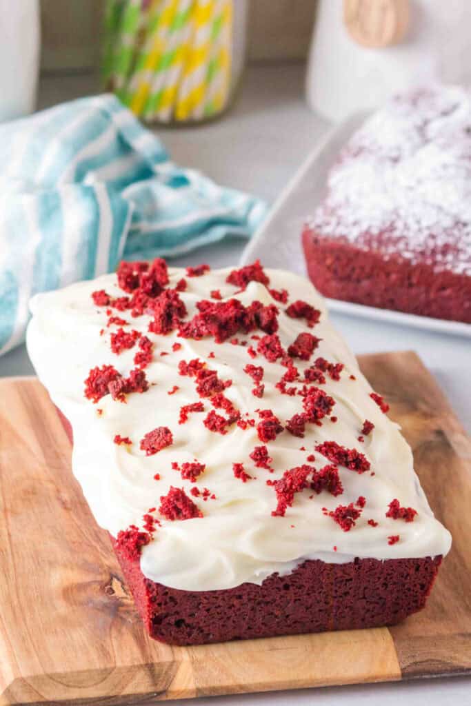 Red velvet loaf topped with thick cream cheese frosting and red cake crumbs on a wooden cutting board.