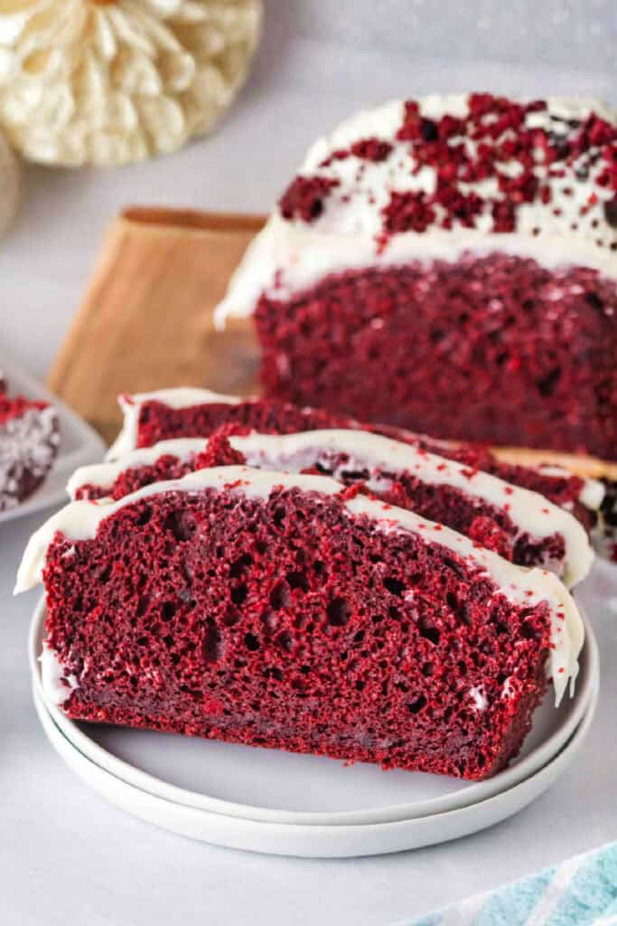 Close-up of a slice of red velvet loaf with cream cheese frosting on a white plate, with the remaining loaf in the background.