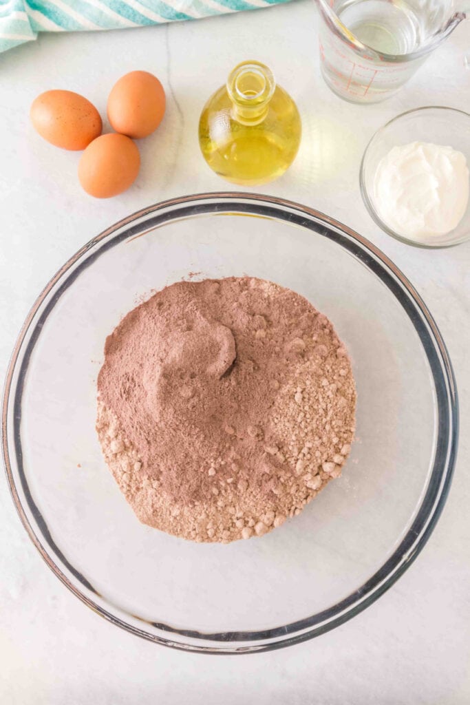 Glass mixing bowl with red velvet cake mix and chocolate pudding mix on a white countertop with eggs, oil, sour cream, and water nearby.