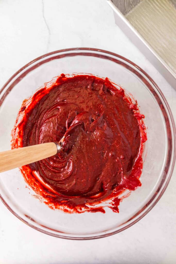 Glass mixing bowl filled with bright red velvet batter being stirred with a spatula.