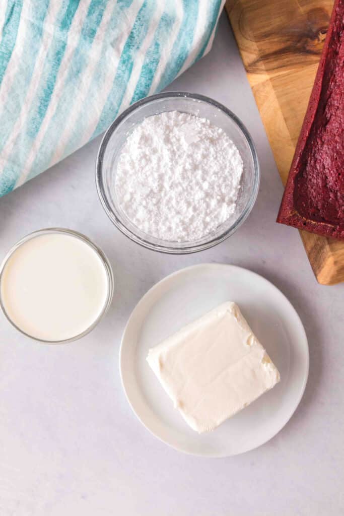 Cream cheese, powdered sugar, and heavy cream arranged in small bowls next to a baked red velvet loaf on a cutting board.