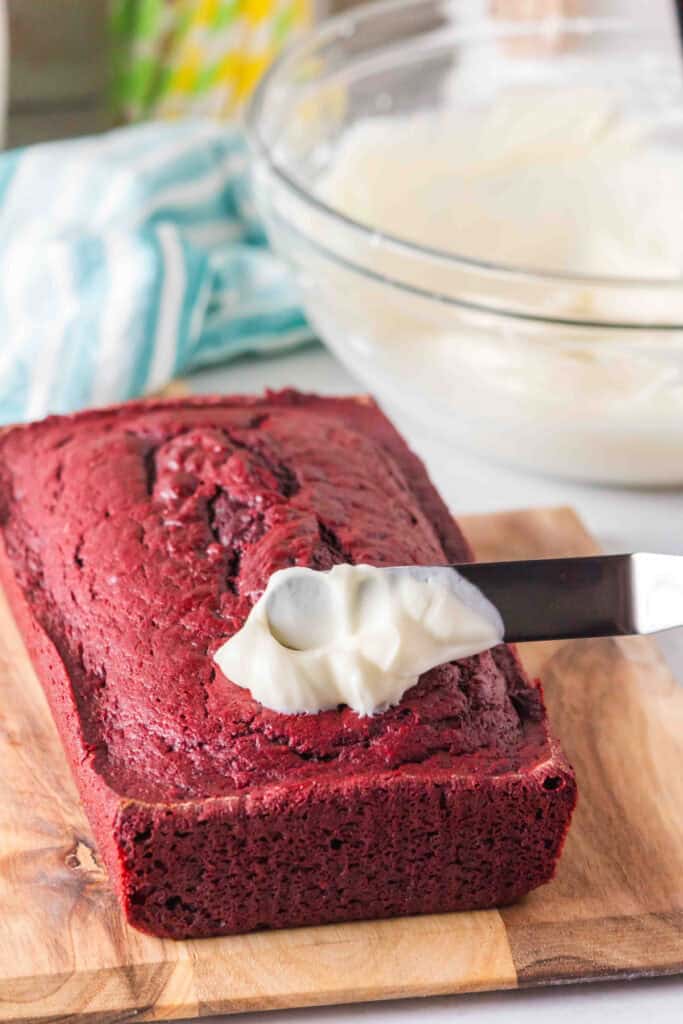 Offset spatula spreading cream cheese frosting over the top of a baked red velvet loaf on a wooden cutting board.