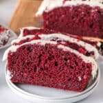 Square image of sliced red velvet loaf with cream cheese frosting on a plate, showing the red interior and white frosting.